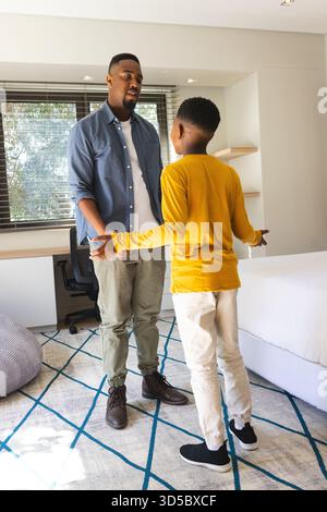Père afro-américain et fils discutant dans une chambre moderne avec lit, tapis gris et chaise de bureau Banque D'Images