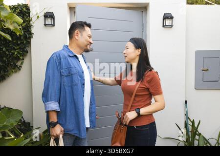 Couple asiatique mature souriant et discutant tout en transportant des sacs à provisions à l'entrée de la maison Banque D'Images