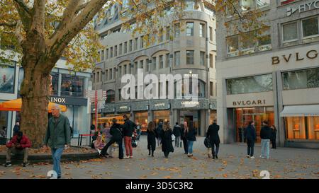 Les gens marchent à travers une place animée de la ville bordée de boutiques tandis que les feuilles tombent des arbres au-dessus. Banque D'Images