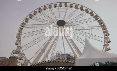 Une grande roue domine un espace de festival, créant une atmosphère animée dans le paysage urbain. Banque D'Images