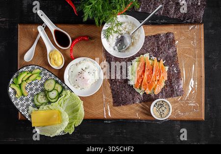 Le processus de fabrication d'un burger sushi créatif avec du saumon, de l'œuf frit, du fromage, du concombre et de l'avocat sur un fond texturé. Étape 4. Vue de dessus Banque D'Images