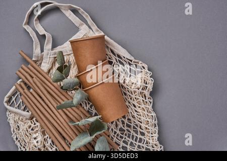 Boire des tubes bruns pailles en papier et fécule de maïs, sac de marché en maille et tasses à café en papier vides sur un fond gris à la mode. Zéro déchet et concept sans plastique. Vue de dessus. Banque D'Images