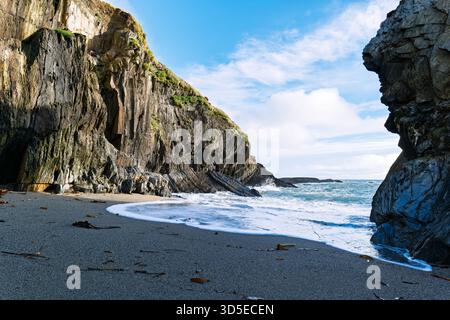 Les vagues s'écrasent sur la plage de sable sombre de Sheep Cove en Irlande, avec des falaises abruptes et rocheuses s'élevant de chaque côté sous un ciel bleu partiellement nuageux. Banque D'Images