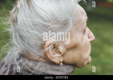 Femme âgée 80-90 ans avec un appareil auditif dans son oreille, gros plan de la personne âgée aux cheveux gris à l'extérieur, concept de vieillissement et de soins de santé Banque D'Images