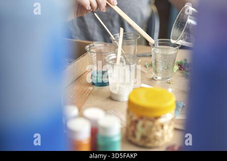 Mains versant de la résine dans des gobelets transparents pour le projet d'artisanat sur la table de travail avec de la peinture et des matériaux Banque D'Images