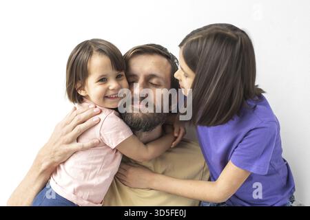 Père embrassant deux filles souriantes, l'une en rose et l'autre en violet, dans un moment familial tendre sur fond blanc Banque D'Images