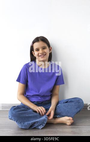 Fille brune blanche souriante âgée de 7-8 ans en t-shirt violet et jeans assis pieds nus sur plancher de bois contre mur blanc Banque D'Images