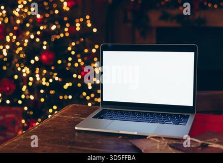 Ordinateur portable avec une maquette d'écran blanc placé sur une table en bois sombre à l'intérieur sur un fond d'arbre de Noël floue et lumineux. Banque D'Images