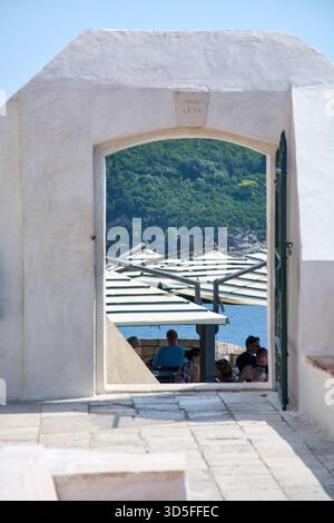 Dubrovnik. Croatie - 15 novembre 2025 : vue à travers une entrée en pierre de Buza Bar, avec des personnes assises sous des parasols surplombant la mer Adriatique an Banque D'Images
