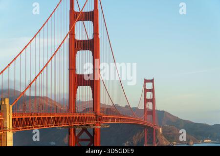 Les tours du Golden Gate Bridge orange vif et les câbles suspendus dominent le cadre contre un ciel clair de San Francisco et des collines lointaines. Banque D'Images