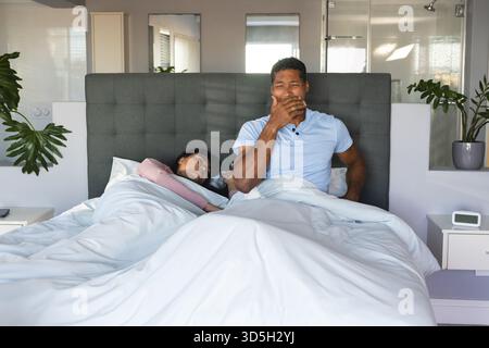 Couple se réveillant dans le lit, homme bâillant tandis que femme dormant paisiblement Banque D'Images