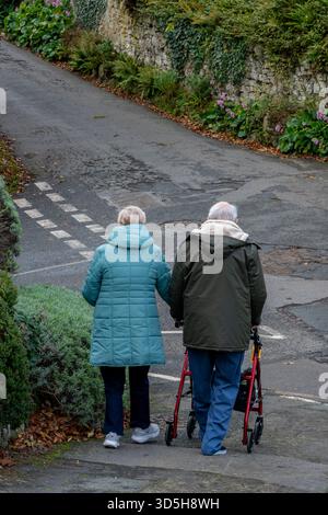 Couple âgé marchant bras dans bras le long d'une route de campagne portant des vêtements chauds et des manteaux d'hiver. Homme utilisant l'aide à la marche assisté par femme dehors pour une promenade. Banque D'Images