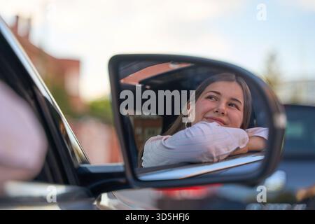 Jeune femme réfléchissant dans un miroir de voiture, regardant vers le haut réfléchi. Elle éprouve la contemplation et la liberté lors d'un Road trip Banque D'Images