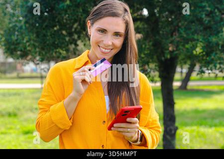 Jeune femme joyeuse debout dans le parc, souriant à la caméra tout en tenant un smartphone et une carte de crédit, représentant les achats en ligne, les services bancaires mobiles, le chiffre Banque D'Images