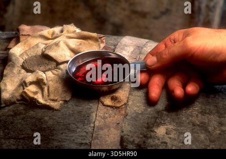 A l’intérieur d’une cave, une main tient une petite tasse en métal remplie de vin rouge, tirée d’un tonneau en bois. France Banque D'Images