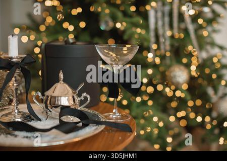 Gros plan d'une table élégante avec un coffret cadeau, de l'argenterie et une coupe de champagne devant un sapin de Noël illuminé Banque D'Images