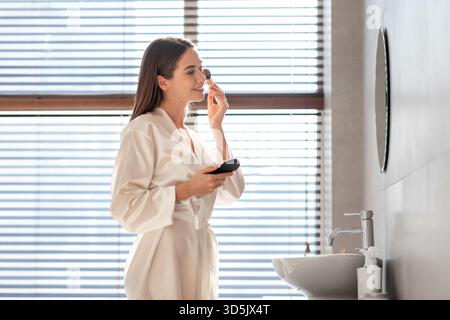Attrayant jeune femme en robe de soie blanche appliquant le maquillage dans la salle de bains de luxe Banque D'Images