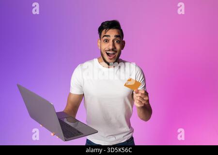Excité jeune homme arabe célèbre la victoire des achats en ligne avec ordinateur portable et carte de crédit Banque D'Images