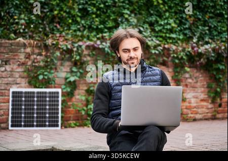 L'homme est assis à l'extérieur, travaillant sur un ordinateur portable sur fond de murs de briques recouverts de lierre. Le panneau solaire repose à proximité, mettant en valeur un espace de travail durable et écologique dans un environnement urbain naturel et serein. Banque D'Images