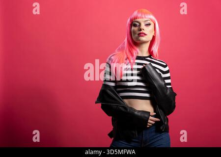 Modèle de mode confiant pose librement, mélangeant élégance et flair punk pour exprimer la beauté rebelle moderne. Jeune femme avec des cheveux roses audacieux roches veste en cuir edgy look dans un portrait de studio élégant. Banque D'Images