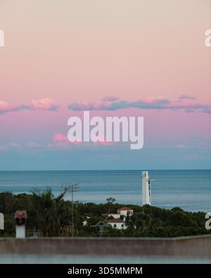 Doux coucher de soleil pastel sur la Méditerranée avec une tour côtière, des toits et une mer calme sous des nuages roses éclatants. Banque D'Images