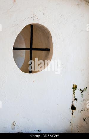 Une fenêtre ronde dans un mur en plâtre blanc présente un motif de croix noire divisant son intérieur. Banque D'Images