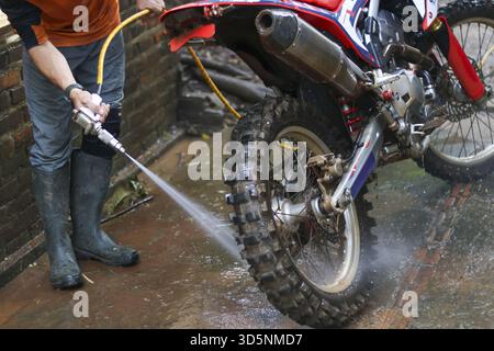 Homme concentré dans des bottes en caoutchouc lavage sous pression moto rouge boueuse. Nettoyage d'entretien avec nettoyeur haute pression sur roue sale de vélo sale après la course Banque D'Images