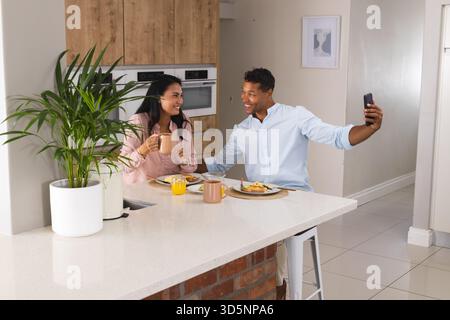 Couple appréciant le petit déjeuner à la maison, prenant selfie avec smartphone, souriant joyeusement Banque D'Images