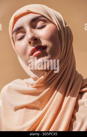 Femme en hijab léger, les yeux fermés, pose sur fond chaud, exsudant le calme et la confiance, mettant en valeur la beauté culturelle et l'atmosphère sereine Banque D'Images