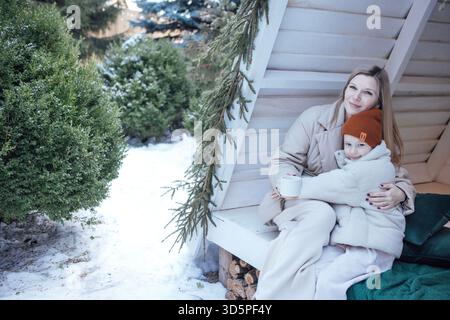 Scène hivernale confortable mettant en vedette femme et enfant enveloppés dans des vêtements chauds, assis sur un porche avec des arbres couverts de neige, partageant une boisson chaude et mome joyeux Banque D'Images