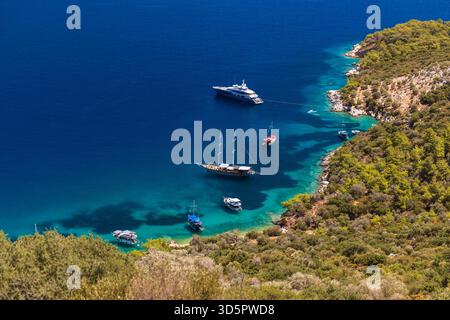 Vue aérienne d'une baie tranquille où plusieurs bateaux et yachts reposent près d'une côte rocheuse parsemée de pins. L'eau turquoise claire contraste avec le bleu profond Banque D'Images