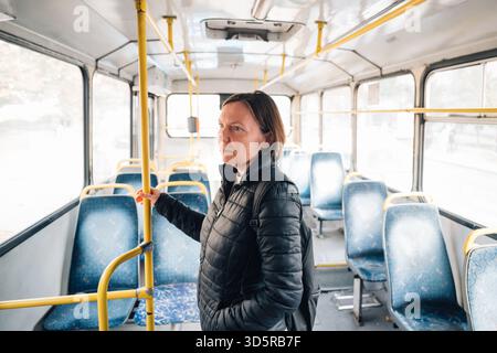 Femme d'âge moyen dans un bus urbain délabré, debout près de la porte et tenant la main courante, symbolisant les déplacements quotidiens en Europe de l'est. Mise au point sélective. Banque D'Images