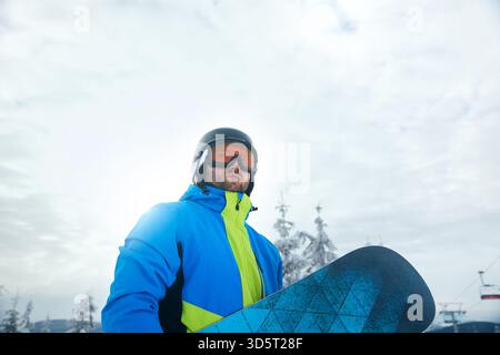 Un snowboarder se tient en toute confiance dans une station de ski, portant une veste colorée et un casque, prêt à dévaler les pistes par une journée neigeuse sous un ciel nuageux. Banque D'Images