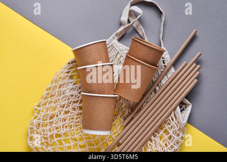 Boire des tubes bruns pailles en papier et fécule de maïs, sac de marché en maille et tasses à café en papier vides sur un fond gris et jaune tendance. Zéro déchet et concept sans plastique. Vue de dessus. Banque D'Images