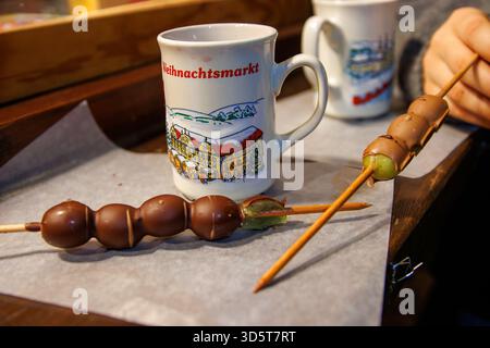 Würzburg, Allemagne, 23 décembre 2024. Brochettes de fruits recouvertes de chocolat placées sur du parchemin à côté de tasses en céramique dans un étal de noël allemand. Banque D'Images