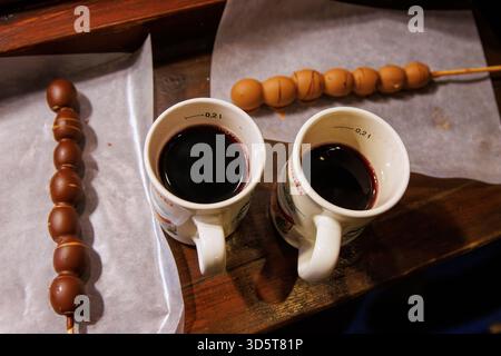 Würzburg, Allemagne, 23 décembre 2024. Deux tasses de vin chaud accompagnées de brochettes de fruits enrobées de chocolat sur papier parchemin lors d'un marché de noël. Haut v Banque D'Images