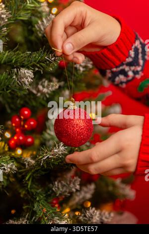 Enfant décorant un arbre de Noël avec un ornement rouge, créant un moment de vacances chaleureux et festif sur un fond rouge vif Banque D'Images