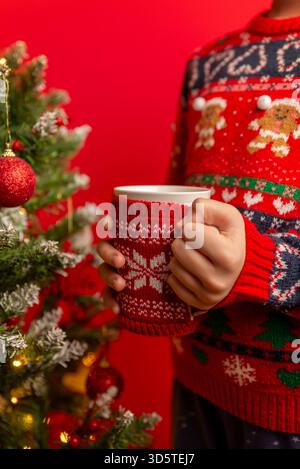 Enfant tenant une tasse de vacances devant un arbre de Noël décoré avec des lumières et des ornements rouges sur un fond lumineux gfh Banque D'Images
