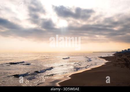 Coucher de soleil doré sur la plage tranquille de Gandia avec vagues, nuages et horizon lointain de la ville Banque D'Images
