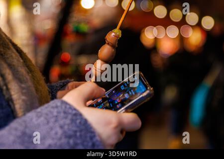 Würzburg, Allemagne, 23 décembre 2024. Jeune femme tenant un smartphone et une brochette de fruits enrobée de chocolat à un marché de noël. Lumières festives chez bl Banque D'Images