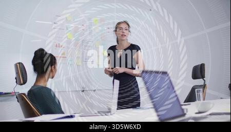 Présentant femme en robe marine faisant des gestes au mur de planification par table de conférence, ordinateur portable ouvert à proximité Banque D'Images