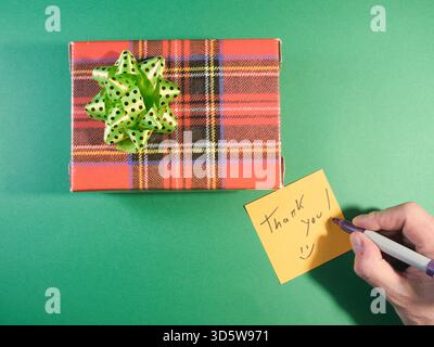 Boîte cadeau de remerciement avec note manuscrite - vue de dessus d'une boîte cadeau festive à carreaux avec un noeud vert et une main écrivant un « Merci ! » note sur un jaune Banque D'Images
