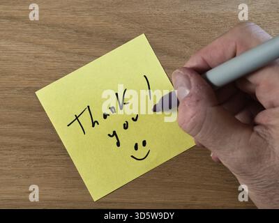 Note de remerciement sur le bureau en bois - gros plan d'une main écrivant « Merci ! » avec un marqueur sur une note collante jaune, placé sur un bureau en bois texturé. Expre Banque D'Images