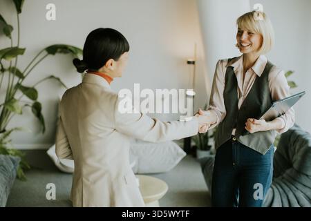 Dans un bureau élégant, deux femmes d'affaires partagent une poignée de main amicale tout en souriant. L'un porte un blazer et tient un ordinateur portable, tandis que l'autre arbore une lumière Banque D'Images