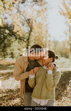 Un couple partage un moment ludique, entouré de feuilles d’automne dorées dans un parc pittoresque, rayonnant de bonheur et de chaleur. Banque D'Images