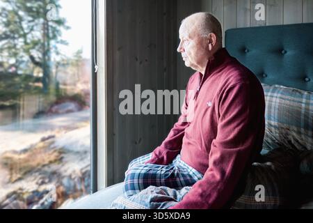Réflexion matinale comme un homme plus âgé est assis près de la fenêtre dans une cabine nordique calme, regardant dehors avec une expression réfléchie. Lumière naturelle douce, paix Banque D'Images
