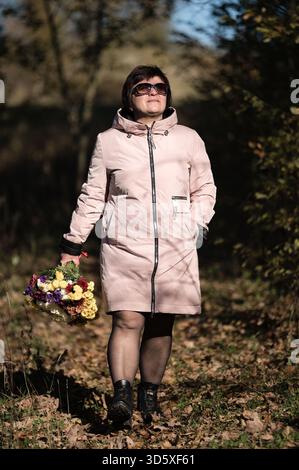 Une femme joyeuse se promène dans une forêt pittoresque, entourée de feuilles tombées, se délectant de la beauté de la nature tout en portant une fleur colorée bo Banque D'Images