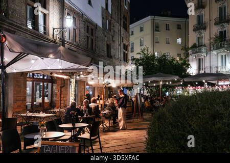 Vue nocturne d'un restaurant animé en plein air à Turin, Italie, avec des gens dînant sous des lumières et des parasols le long d'une charmante rue historique. Banque D'Images