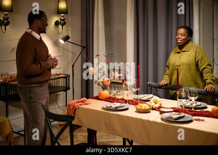 Homme noir et femme noire debout près de la table à manger décorée préparant le dîner de Thanksgiving entre amis, à la fois souriant et engageant dans la conversation, nourriture de fête et bougies visibles Banque D'Images