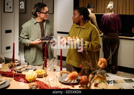 Groupe multiethnique de jeunes adultes debout autour de la table partageant de la nourriture pendant le dîner de Thanksgiving, homme caucasien tenant une plaque de raisins parlant à une femme noire tenant une plaque de muffins Banque D'Images